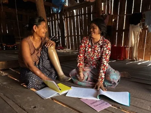 Two women sitting in a house, they have documents in front of them. They are smiling at each other.