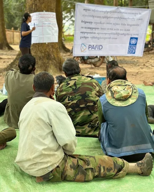 A group of men from behind, seated on the gorund, listening to a training outside. A woman in the back is standing and speaking.