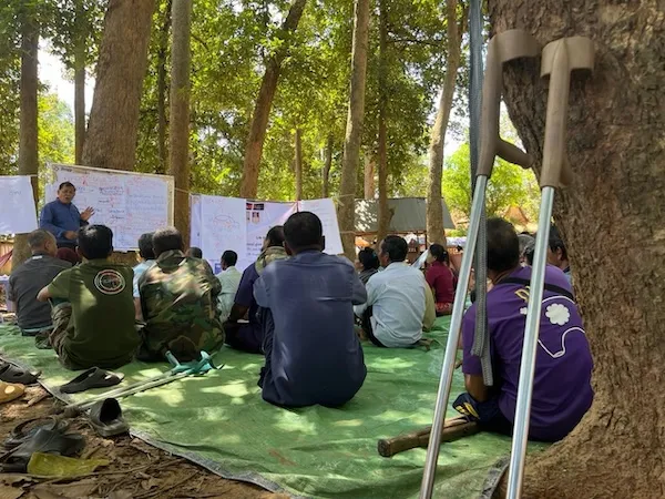 Group of people seated outside, following a training. Crutches have been left next to a tree.