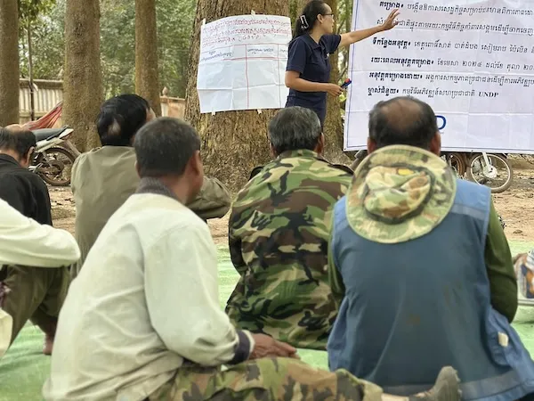 A group of men from behind, seated on the gorund, listening to a training outside. A woman in the back is standing and speaking.
