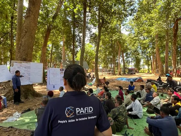 Group of people attending a training. They are seated outside. A woman who's back is facing us wears PAfID's shirt.