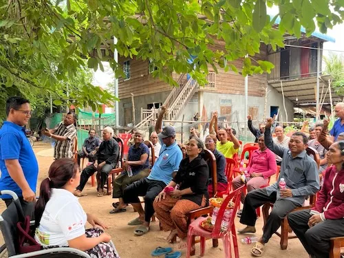A group of people of sitted outside, under some trees, they are attending a meeting, raising hands to participate.