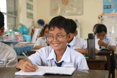 Mr. Ket Monireach smiling. He is seated in his classromm and is writing in his notebook.