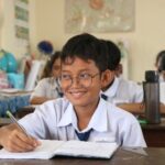 Mr. Ket Monireach smiling. He is seated in his classromm and is writing in his notebook.