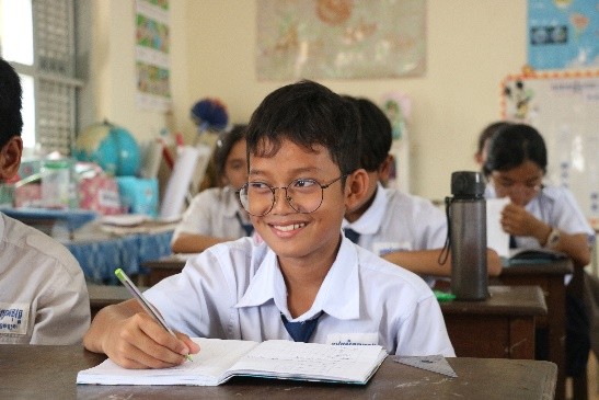 Mr. Ket Monireach smiling. He is seated in his classromm and is writing in his notebook.